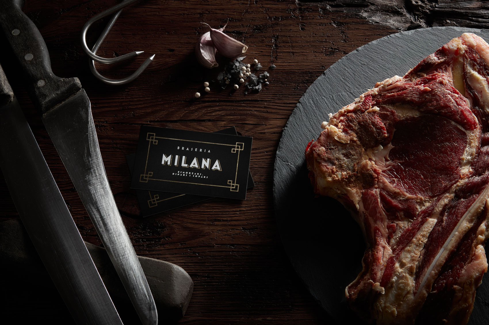 Business card with new logo of restaurant grill Braseria La Milana next to t-bone steak and knife